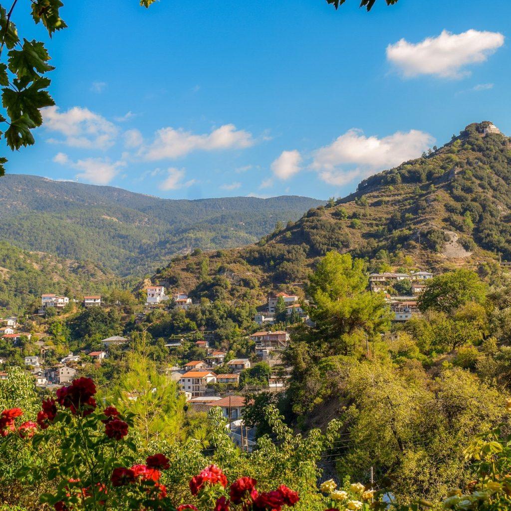 View of a mountain village at the foot of a hill, with blue skies and greenery surrounding. Troodos, Cyprus. Presented by Comark Estates.