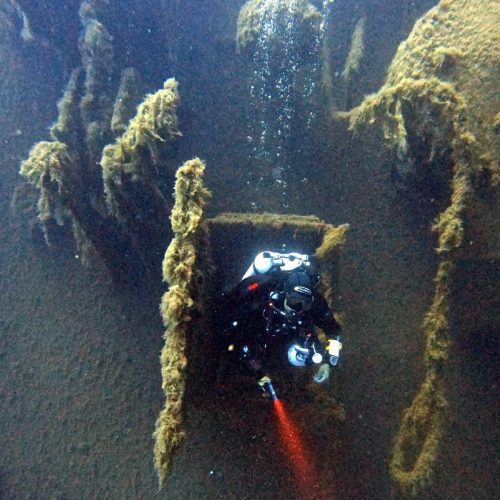 Diver emerging from shipwreck door underwater, holding red beam light. Zenobia Shipwreck Cyprus.